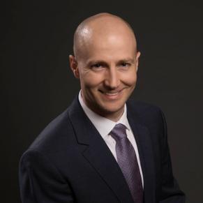 A man in a black suit, gray tie, and white collared shirt stands and smiles