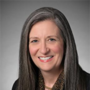 A woman with long gray hair and a black shirt stands and smiles