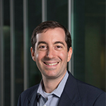 A man in a blue suit and light blue collared shirt stands and smiles