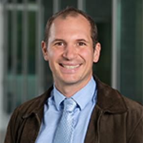 A man in a brown jacket, blue collared shirt, and silver tie stands outside and smiles