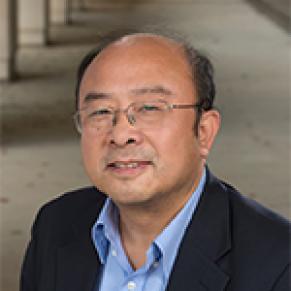 A man in a black suit and blue collared shirt stands and smiles