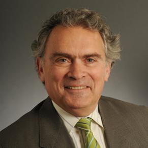 A man in a brown suit with a green tie and a collared shirt stands smiling